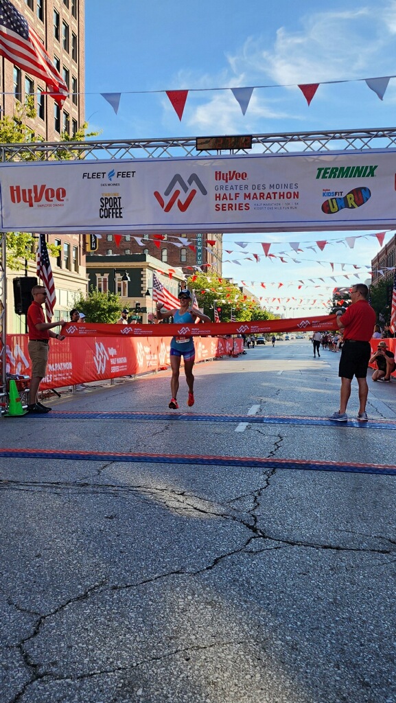 Runner crossing the finish line at a marathon event in a city street, with banners overhead and people watching nearby.